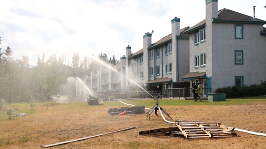 Des maisons à droite, avec des gicleurs à grand débit projetant de l’eau vers les arbres à gauche.