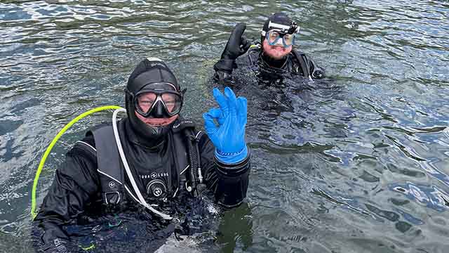 Deux personnes vêtues d’un équipement de plongée sous-marine saluent la caméra depuis l'eau.