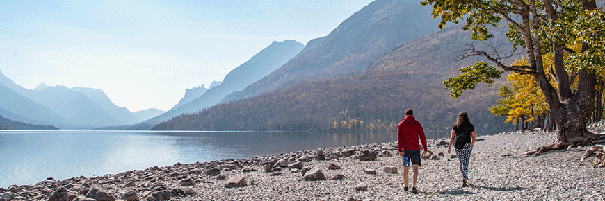 Two people walk along a rocky beach on front of a lake in a valley of mountains. 