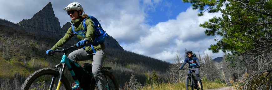 Two mountain bikers descend a path with a mountain in the background.