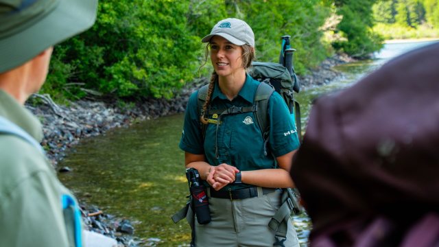 A Parks Canada staff member speaks to a visitor on a guided hike with trees and water in the background
