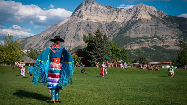 An individual in traditional Blackfoot regalia is seen in a grassy field with Vimy Peak in the background