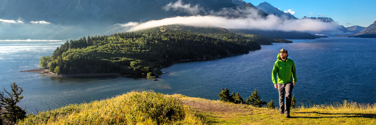 A hiker on the Prince of Wales Hill with views of Upper and Middle Waterton Lakes, hanging clouds and mountain peaks in the background