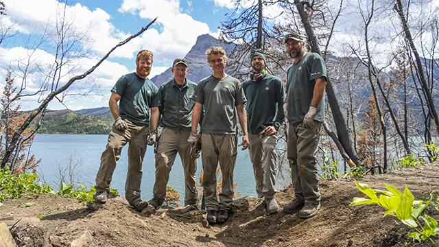 Five people in Parks Canada uniforms stand on a cleared trail on a sunny day.