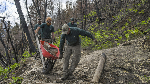 Parks Canada staff move a wheelbarrow of rocks across a damaged section of trail.
