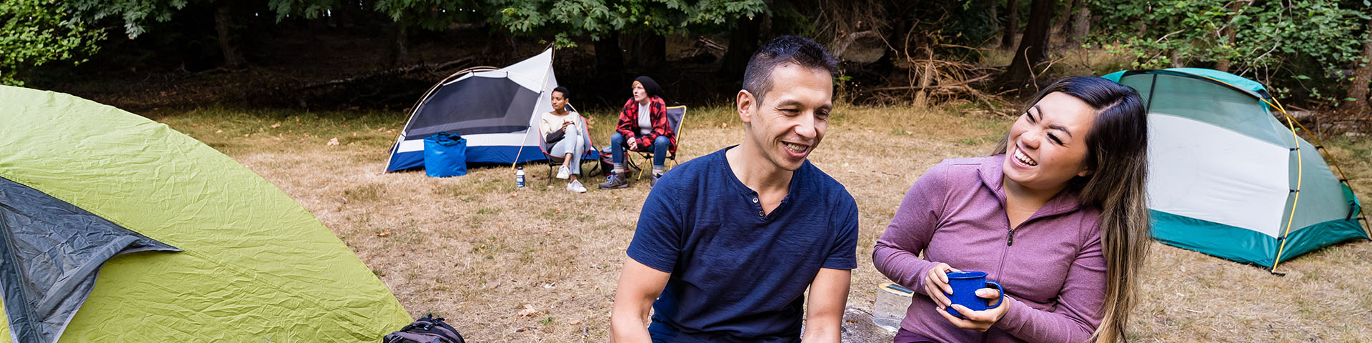 Two individuals are sitting and smiling while enjoying a drink in a mug. In the background, we can see three tents in a wide grassy area with another couple visible.