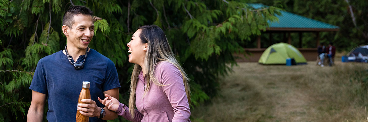  Two people chat while walking on a trail with tents and a picnic shelter in the background.