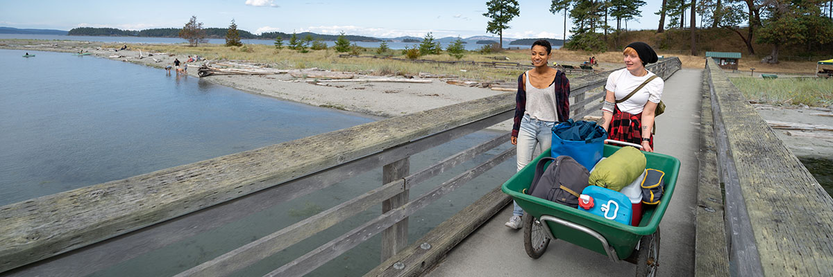 On a dock, two people push a cart filled with camping gear. The beach and sea are visible in the background.