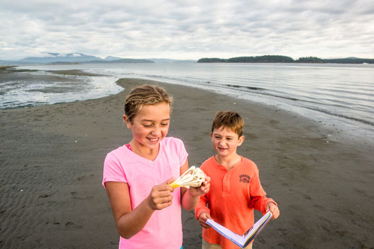 Deux enfants sur une plage et regardant un crâne (accessoire) et le livret Xplorateurs.