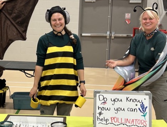 Two women at an educational booth, one dressed in a bee costume, promoting awareness about helping pollinators indoors.
