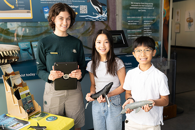 A Parks Canada Interpreter and two children are in an exhibit room at the Shaw Centre for the Salish Sea. The children, holding marine mammal figurines, are all smiling at the camera. 