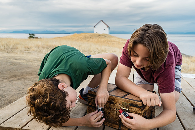 Two kids are kneeling on a wooden platform while trying to open a treasure chest. In the background tall grass and a white building can be seen.