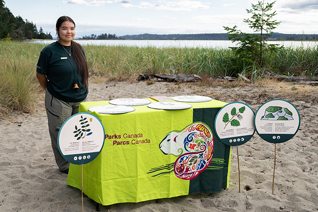 Un interprète de Parcs Canada se tient à côté d’une table d’activités ornée de panneaux représentant des plantes. La plage est visible en arrière-plan. 