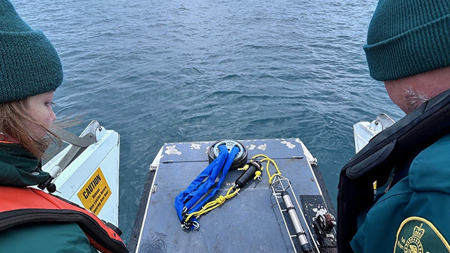 Two people on board a marine vessel observe equipment, with the sea in the background. 