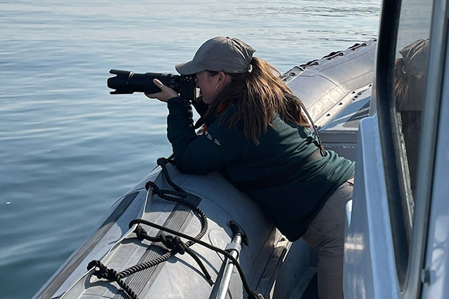 A person using a camera leans on the gunwale of a marine vessel, with the sea in the background.