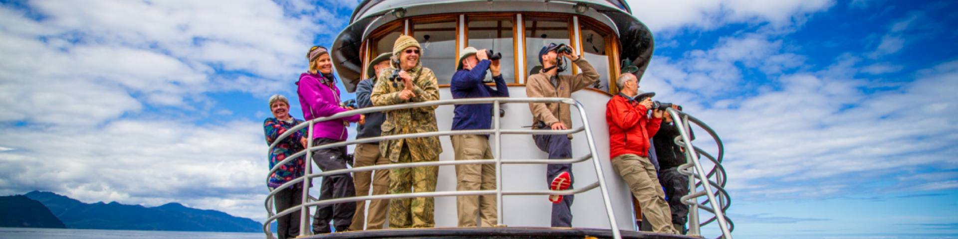 A tour group looks out over the ocean from the bridge of the boat. 3 of them are using binoculars.