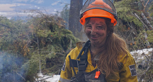 Soot-covered female firefighter standing next to burning brush piles