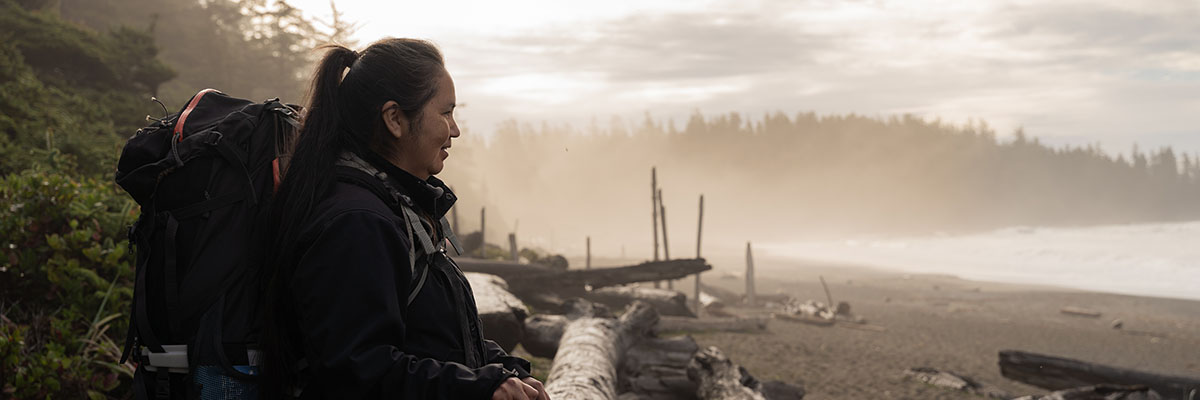 A hiker looks out over the water on a misty day. They are standing on a driftwood-covered beach along the West Coast Trail.