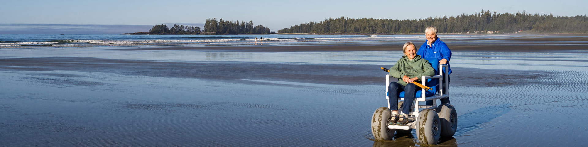 A person in a beach wheel chair with another person pushing the chair on a long sandy beach.