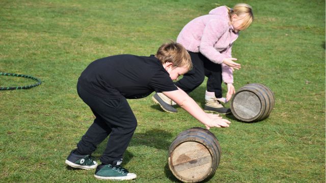 Two children rolling small wooden barrels on grass in a playful competition.
