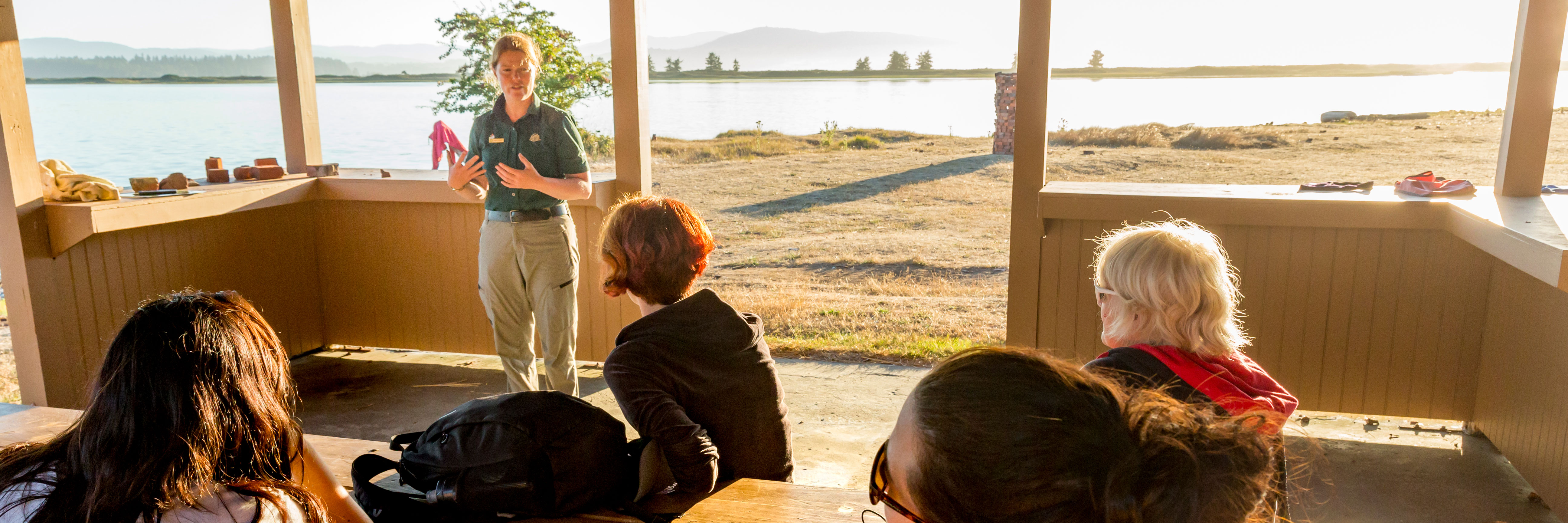 Parks Canada staff member in uniform gestures while giving demonstration to four hikers seated in front of them