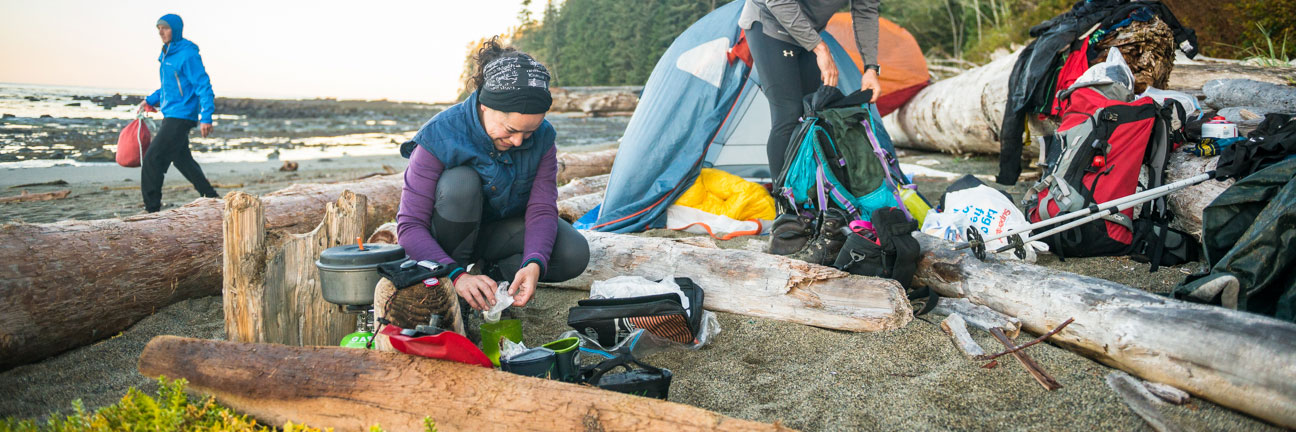 Campers unload packs at beach campsite with gear spread out around them. One makes dinner on camp stove. Two tents set up