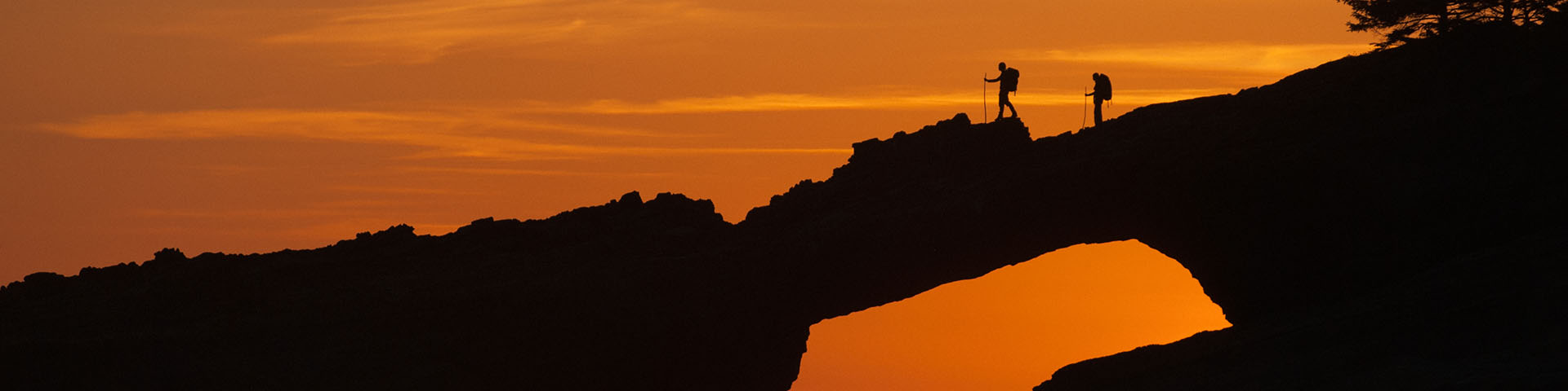 Two hikers in silhouette with packs and poles, cross natural land bridge above water. Orange sunset fills sky in background