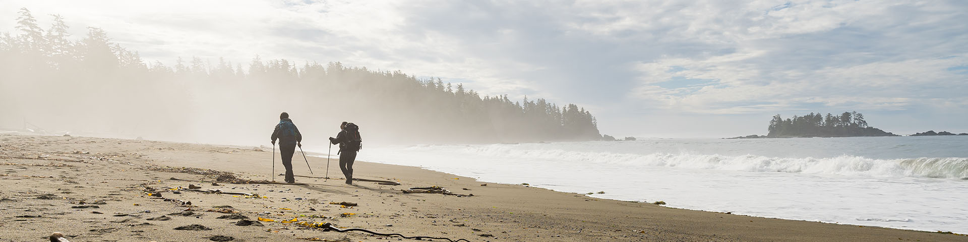 Two hikers with packs hiking along beach with a misty forest in background