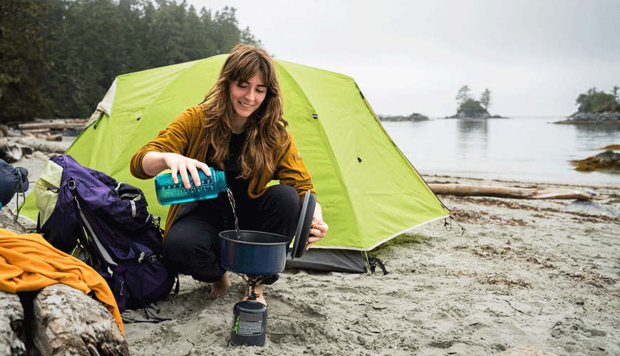 A person cooking on a small camp stove on the beach with a tent and the ocean in the background