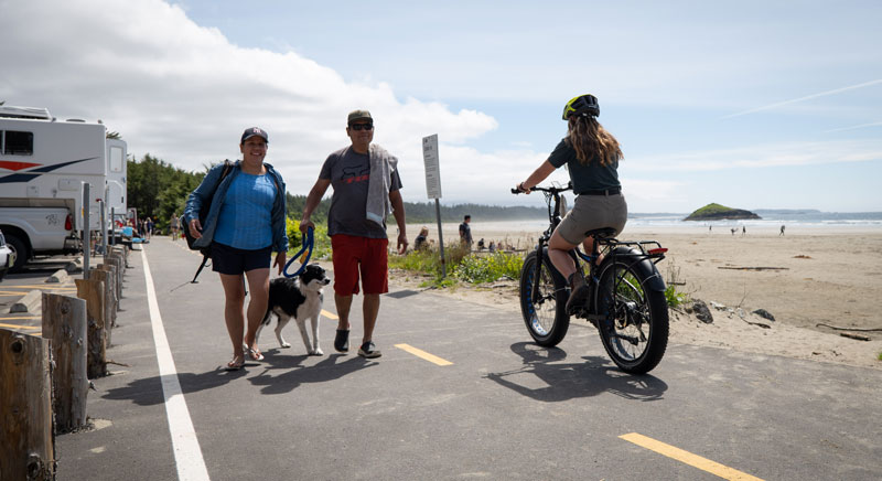 A cyclist riding along right hand side of pathway with couple and dog on leash on left side of pathway. Beach and Lovekin Rock in the background.