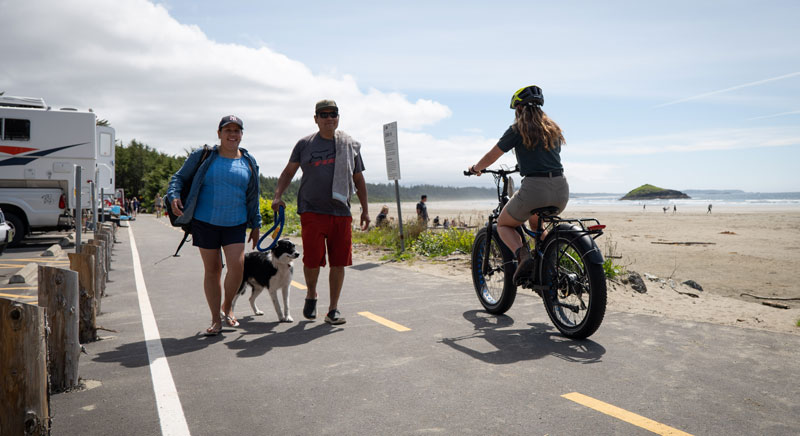Cycliste qui roule sur le côté droit de la piste et couple avec un chien en laisse qui se promène sur le côté gauche de la piste. Plage et Lovekin Rock en arrière-plan.