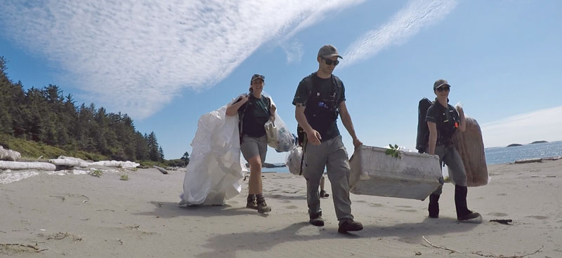 Trois employés de Parcs Canada transportant des débris marins sur une plage de sable.