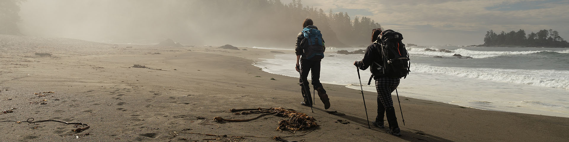 Two hikers with backpacks walking along sandy beach