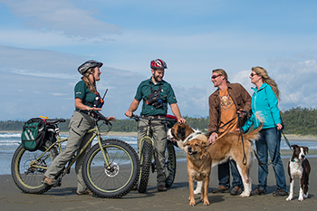 Two Parks Canada staff on bikes talking with visitors and their dogs on leash on the beach