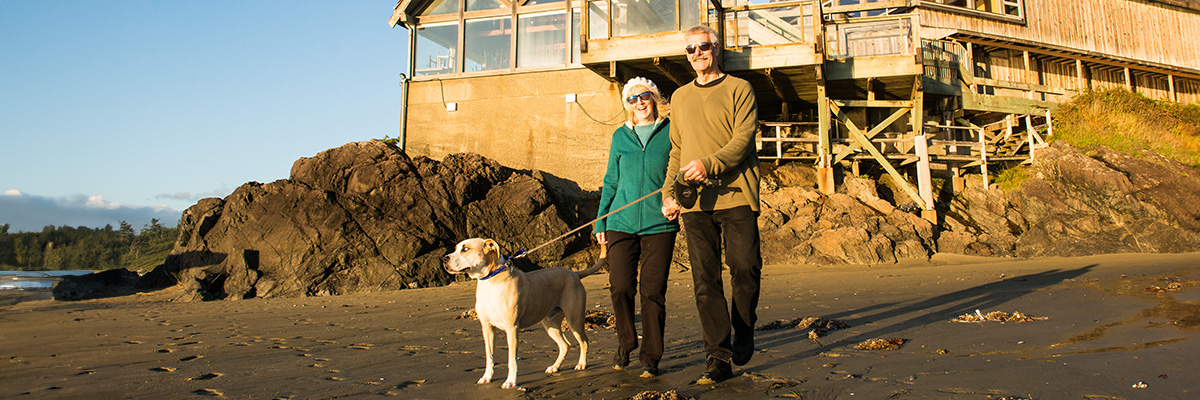 A retired couple enjoy a walk on Wickaninnish Beach with their dog, passing by the Kwisitis Visitor Centre.