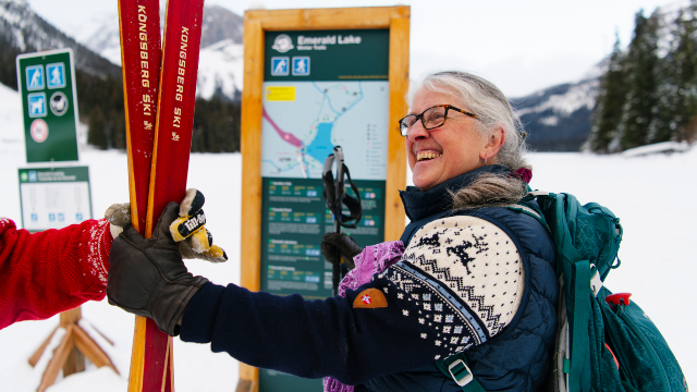 A person smiles at someone who hands them their skis while standing in front of a directional sign at Emerald Lake.