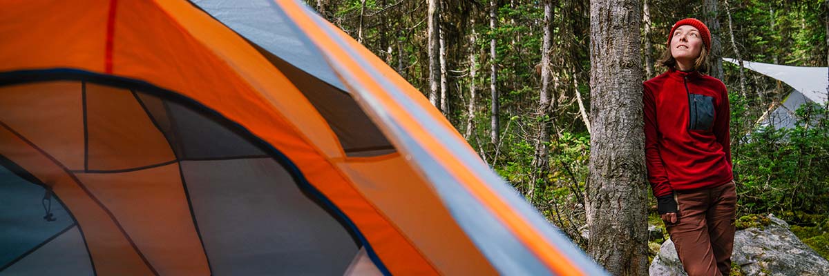 A woman leans on a tree with a tent in the foreground