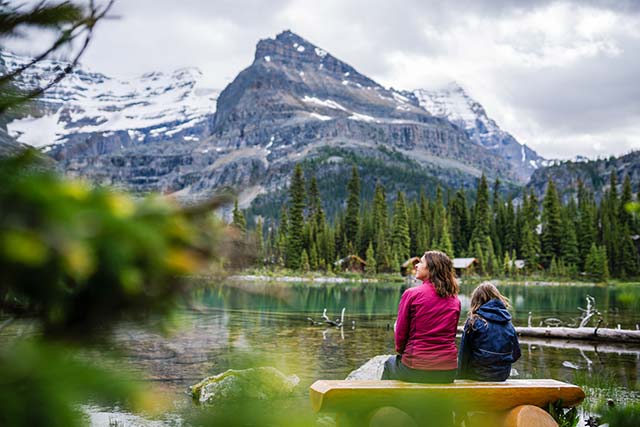 Une femme et une fillette assises sur un banc en rondins au bord du lac O’Hara profitent de la vue sur les montagnes. 