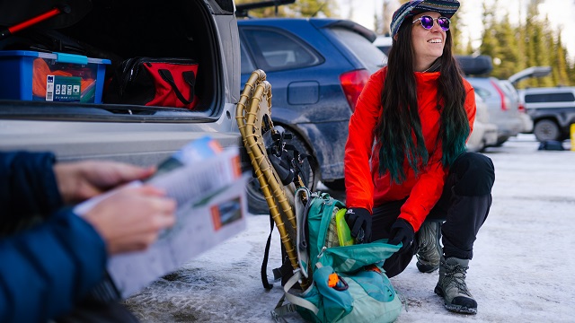 A person, at the back of a car, gets their hiking gear ready (snowshoes, backpack), while another person  looks at a brochure.