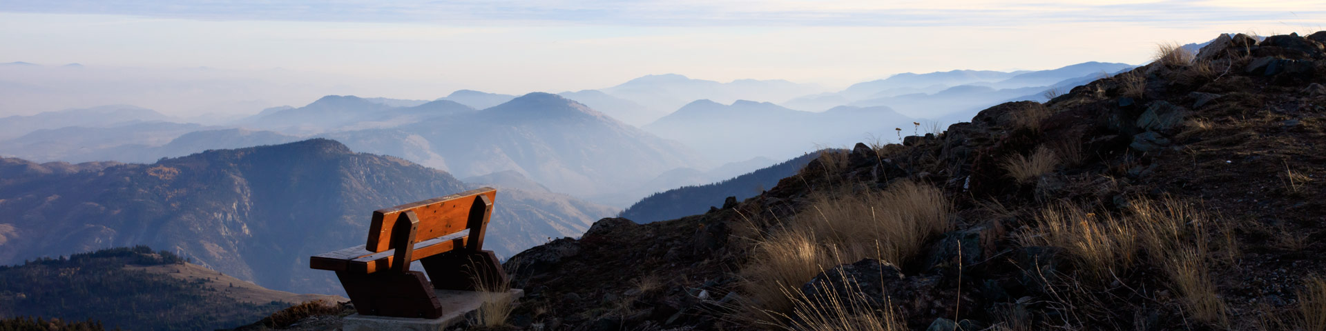 A wooden bench sits on a grassy hilltop. In the distance, mountains stretch for miles and miles.