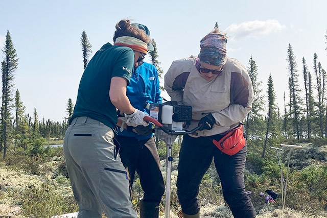 Two people steady a large drill in a forest as a third person watches them.