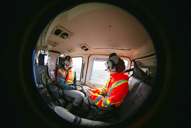 Two people wearing orange safety vests and headsets look out the window of a helicopter.