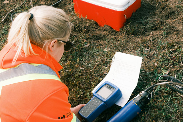 A person in an orange safety vest looks at a piece of monitoring equipment and a sheet of paper resting on soil.