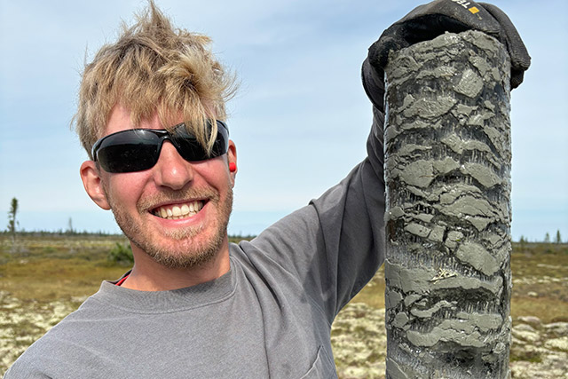 A person wearing sunglasses and smiling holds up a cylinder of soil while standing in a field.