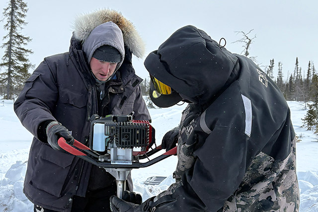 Two people in winter clothing hold onto a large drill in the snow.