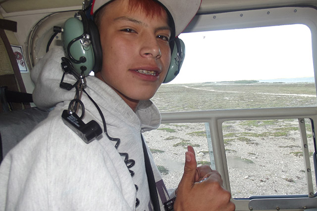 A boy wearing a hat and headphones makes a two-thumbs-up gesture while sitting in a helicopter.