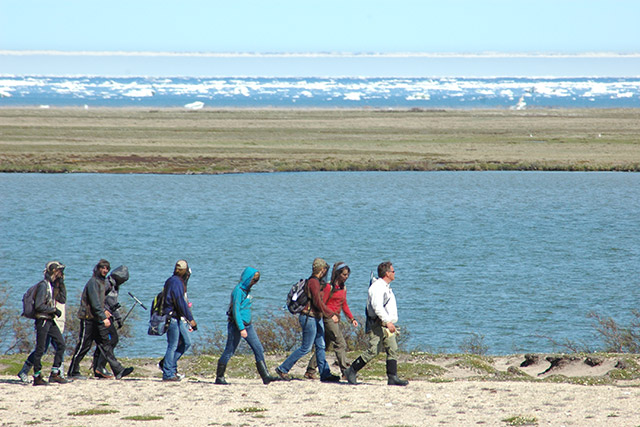 A group of people hikes on sand with a body of water and ice chunks in the background.