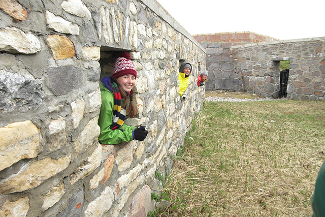 There is a stone wall with three windows and another stone wall in the background. A person is looking at the camera out of each window. 