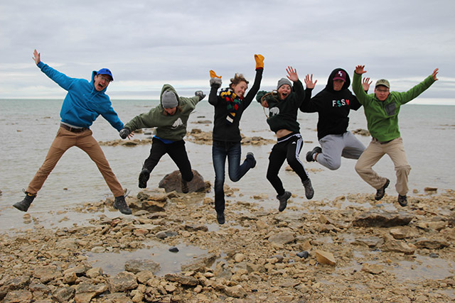 Six people jump in the air. There are rocks and small puddles below them and there is a body of water behind them.