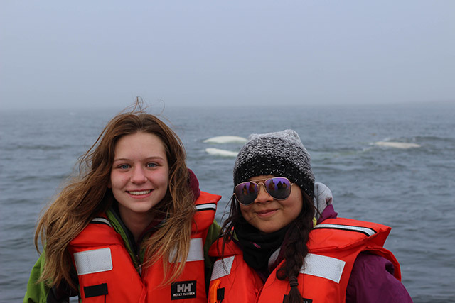 Two people wearing lifejackets smile for a photo. There are three beluga whales swimming in the water in the background.